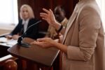 Cropped shot of a female expert witness in beige blazer standing by tribune with microphone and testifying.